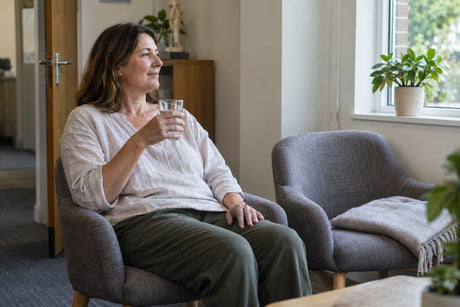 A woman sits back in an armchair in a clinic waiting room, holding a glass of water after an acupuncture session