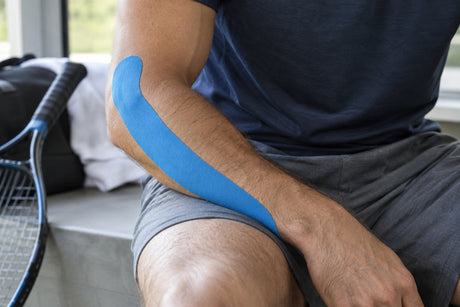 Blue kinesiology tape applied in a single strip along the forearm and outer elbow of a seated man, with a tennis racket beside him