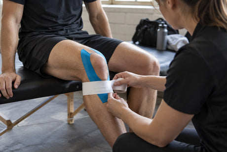 A therapist wrapping white rigid tape over blue kinesiology tape already applied to a seated man's knee