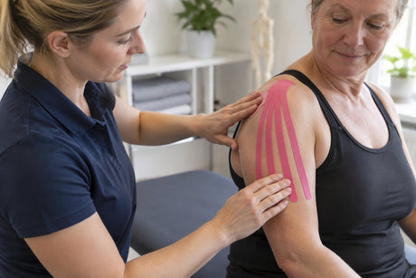 A physiotherapist applying a pink fan of kinesiology tape strips across an older woman's upper arm and shoulder
