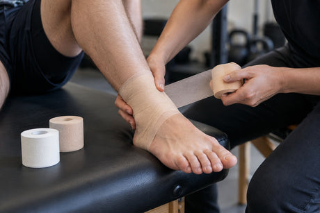 A therapist wrapping beige foam underwrap around a patient's ankle before taping, with rolls of tape on the couch