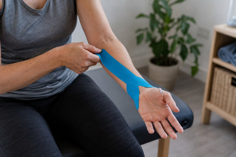 A woman applying a strip of blue kinesiology tape along her forearm towards the wrist while seated
