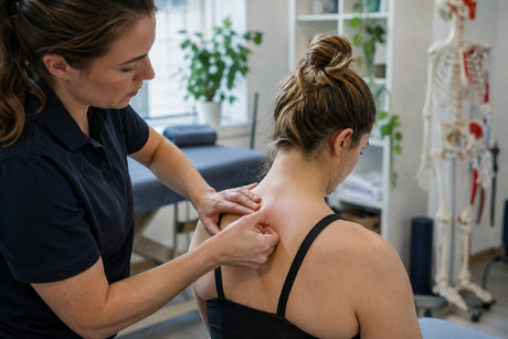 A therapist palpating a seated woman's upper back and shoulder before needling, with a skeleton model behind