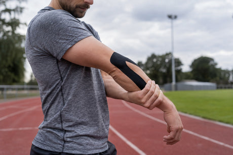 A man on an athletics track holding out his arm to show black kinesiology tape applied around the elbow