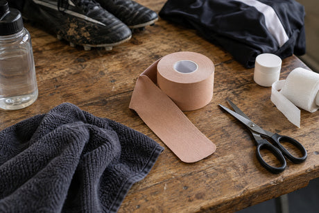 A roll of beige kinesiology tape part unwound on a wooden bench beside scissors, a towel, a water bottle and football boots