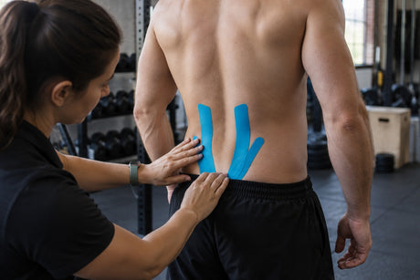 A therapist applying two strips of blue kinesiology tape either side of a standing man's lower spine in a gym