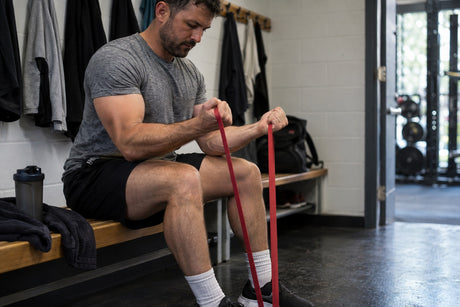 A man sitting on a changing room bench pulling a red resistance band looped under his foot