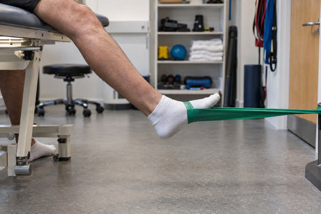 A man sitting on a treatment couch pushing his foot against a green resistance band for an ankle exercise