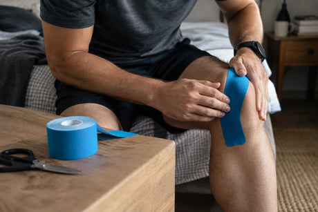 A man sitting on a bed applying a strip of blue kinesiology tape to his knee, with a tape roll and scissors on the table