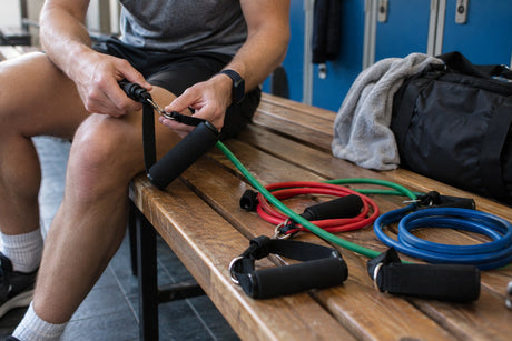 A man clipping a handle onto a resistance tube, with green, red and blue tube bands and handles laid out on a bench