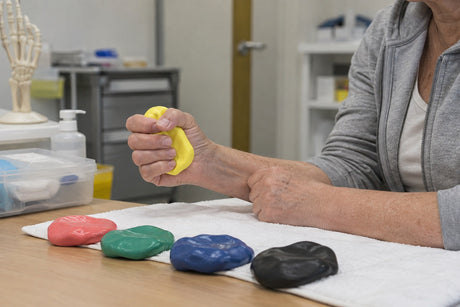 An older woman squeezing yellow therapy putty in a clinic, with red, green, blue and black putty on a towel in front of her