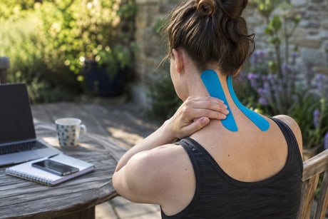 A woman working at an outdoor table reaching back to blue kinesiology tape applied along her neck and shoulder