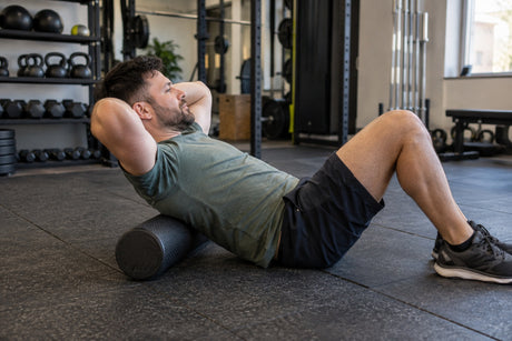 A man lying back over a black foam roller positioned under his upper back in a gym