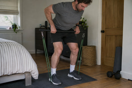 A man in a bedroom performing an upright row on a resistance bar with green bands anchored under his feet