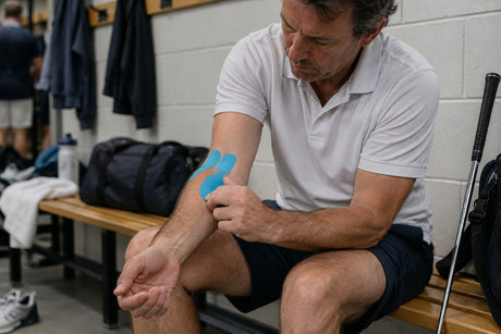 A man on a changing room bench applying blue kinesiology tape to the inside of his elbow, with golf clubs beside him