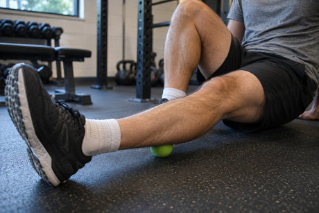 A man sitting on a gym floor rolling the back of his calf over a green massage ball