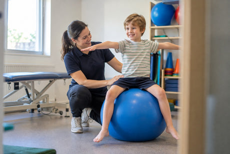 A physiotherapist supporting a young boy balancing with arms outstretched while sitting on a large blue gym ball