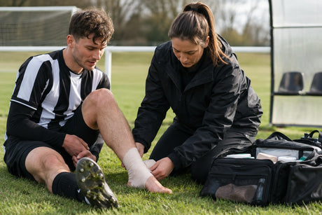 A pitch-side first aider bandaging a footballer's ankle on the grass beside an open medical bag