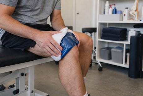 A man sitting on a treatment couch holding a blue gel cold pack wrapped in a towel against his knee