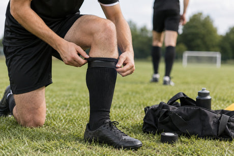 A footballer kneeling on a pitch wrapping black sock tape around the top of his sock, with a roll of tape on the grass