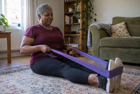 An older woman sitting on the floor at home pulling a purple resistance band looped around both feet