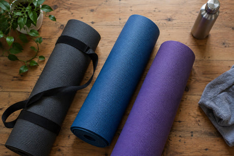Three rolled yoga mats in grey, blue and purple on a wooden floor with a carry strap, water bottle and towel