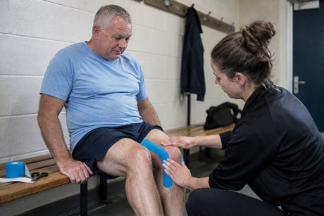 A therapist applying blue kinesiology tape to a seated man's knee in a changing room, with a tape roll and scissors on the bench