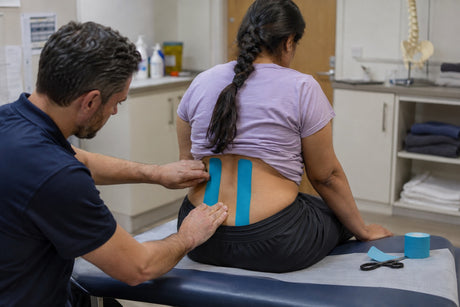 A therapist applying two vertical strips of blue kinesiology tape either side of a seated woman's lower spine