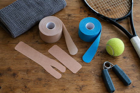 Rolls and cut strips of beige and blue kinesiology tape on a wooden table with a tennis racket, ball and hand gripper