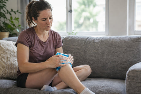 A woman sitting on a sofa holding a blue gel cold pack against her knee