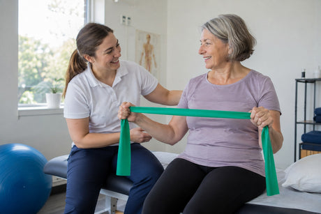 A physiotherapist guiding an older woman pulling a green latex-free resistance band apart while seated on a couch