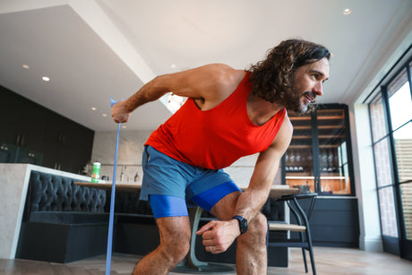 Man in red tank top and blue shorts exercising with resistance bands in a modern kitchen.
